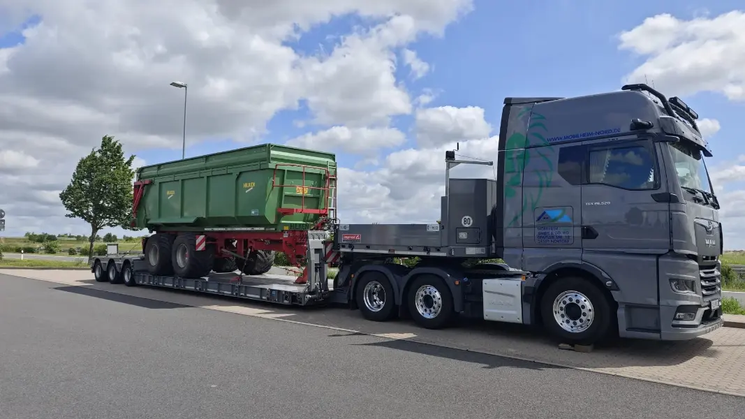 LKW mit einem Tiefbett-Auflieger, auf dem ein großer, landwirtschaftlicher Anhänger verladen ist.