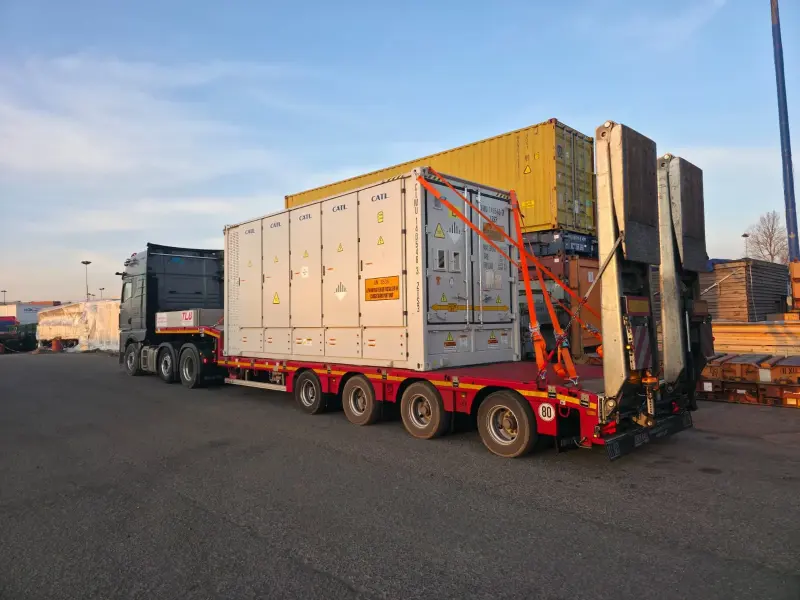 LKW mit einem Semi-Tiefbett-Auflieger, auf dem ein BESS-Container mit Zurrgurten gesichert ist.